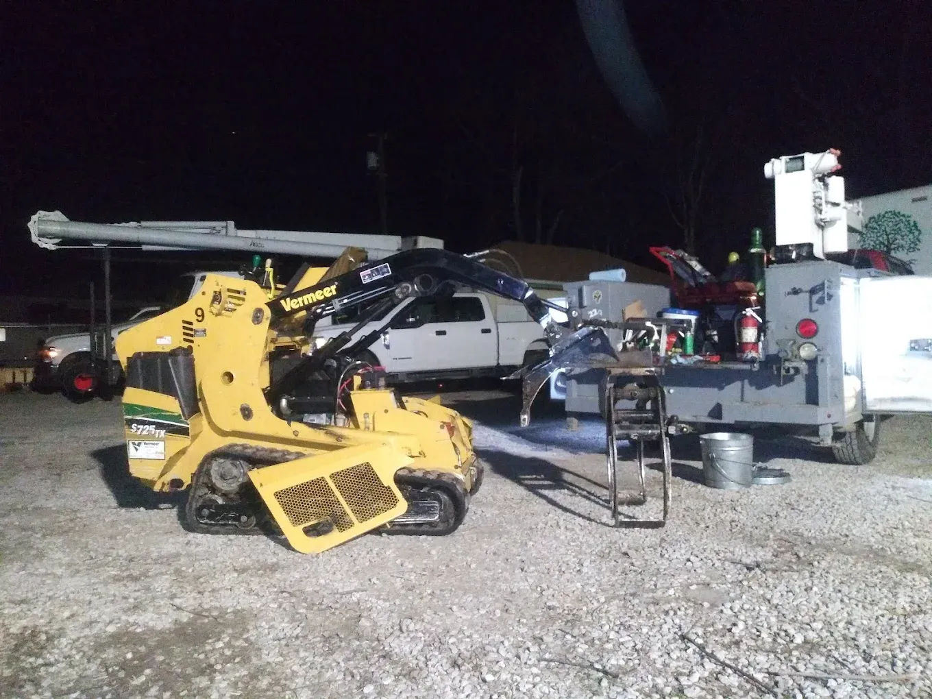 Yellow skid steer loader parked next to a work truck under nighttime lighting